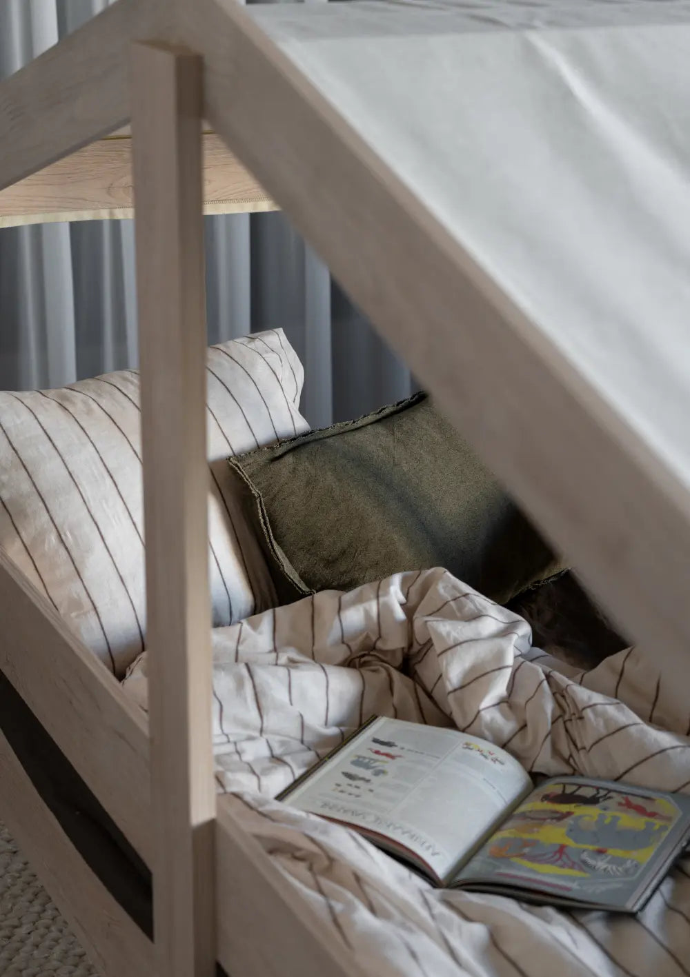 Nahaufnahme der gemütlichen Liegefläche im Quax Hausbett Cottage Natural Ash (200 x 90 cm) mit gestreifter Bettwäsche, einem olivgrünen Kissen und einem aufgeschlagenen Kinderbuch.