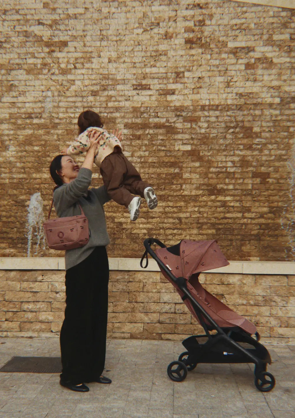 Eine lachende Mutter hebt ihr Kind vor einer Steinwand in die Luft; daneben steht der faltbare Bugaboo Buggy Butterfly 2 x Konges Sløjd Dusty Pink Cherry mit dem passenden Organizer über ihrer Schulter.