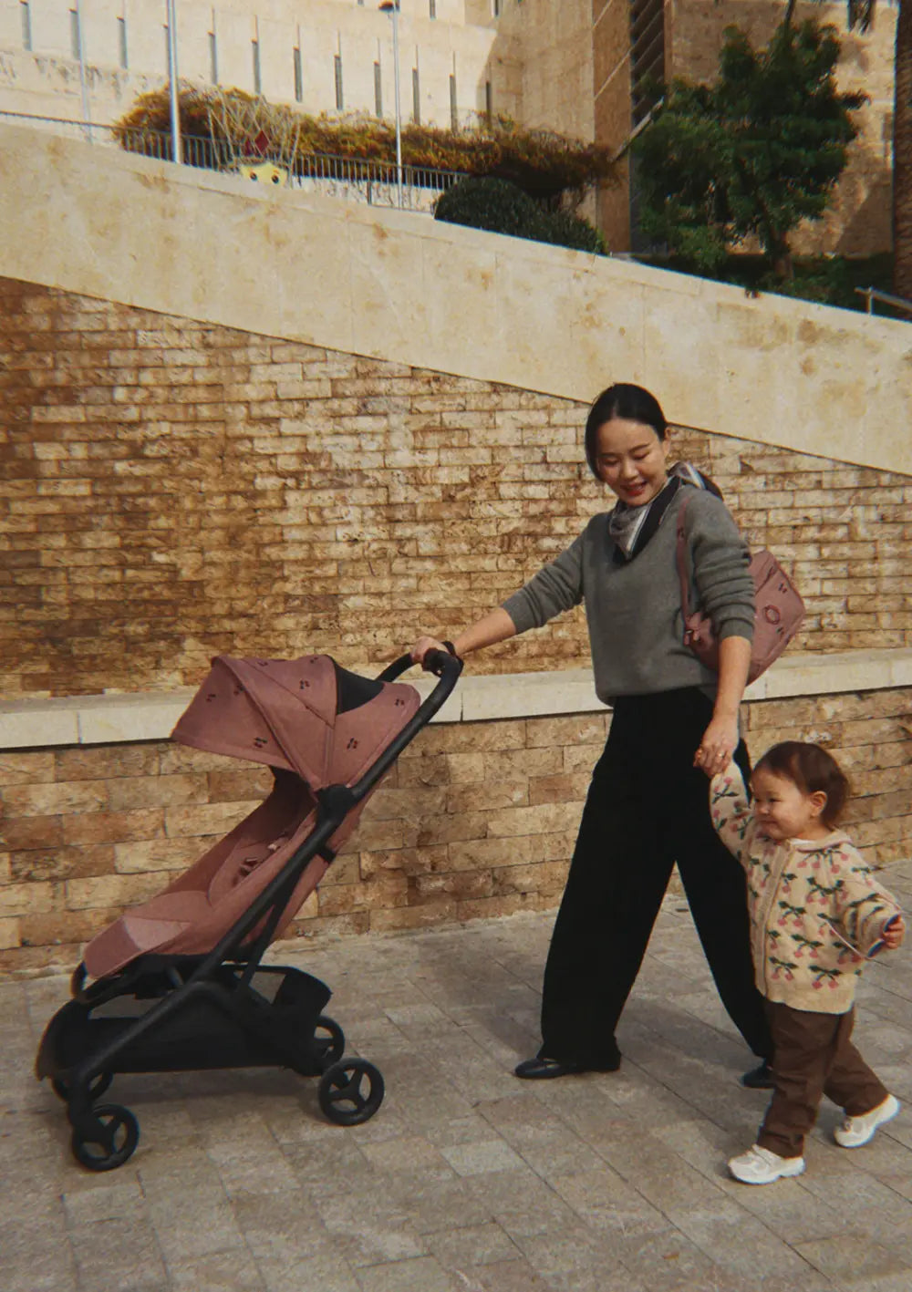 Eine Mutter schiebt den kompakten Bugaboo Buggy Butterfly 2 x Konges Sløjd Dusty Pink Cherry und führt ihr kleines Kind an der Hand; der stylische Reisebuggy besticht durch sein altrosa Kirschmuster-Sonnendach.