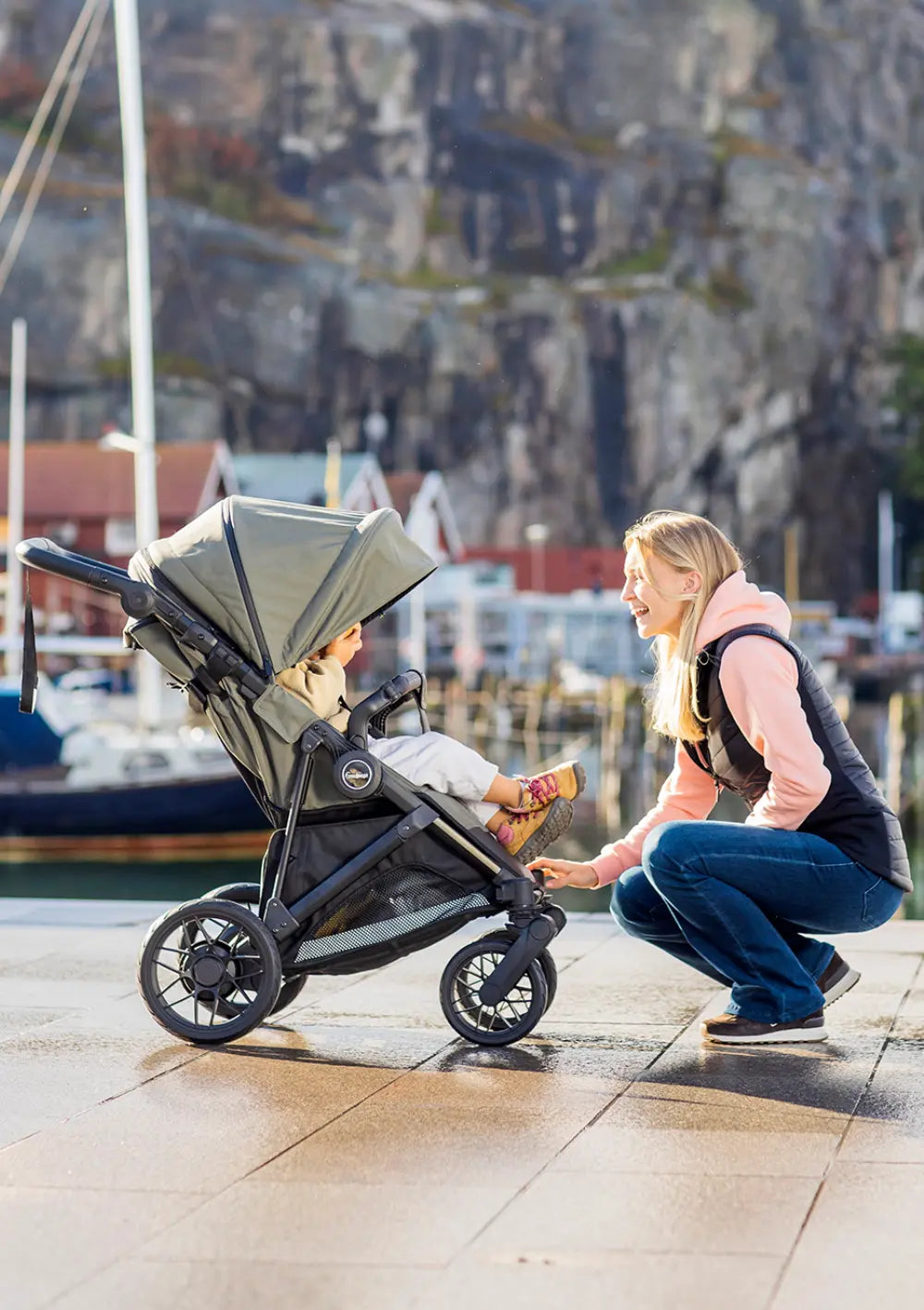 Eine sich hinhockende Mutter lächelt ihr Kleinkind an, das im olivgrünen Emmaljunga Kinderwagen Urban Green an einer Hafenpromenade sitzt.