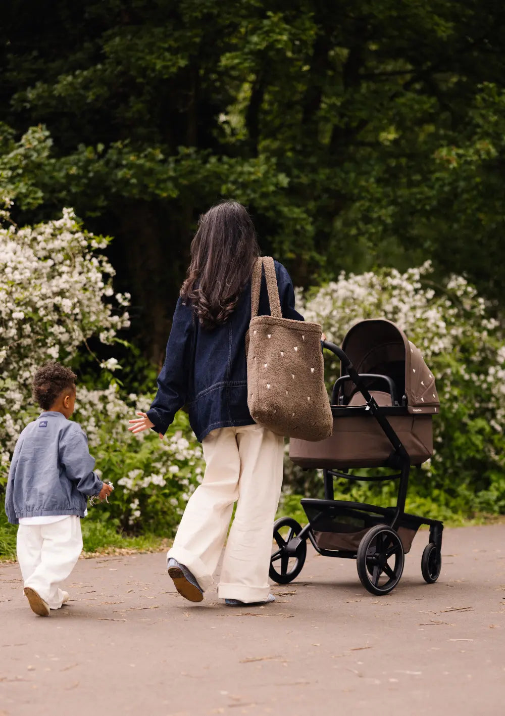 Rückansicht einer Frau mit großer brauner Teddy-Tasche, die den braunen Kombikinderwagen aus dem Joolz Kinderwagen-Set Day5 x Studio Noos schiebt, während ein Kleinkind neben ihr herläuft.