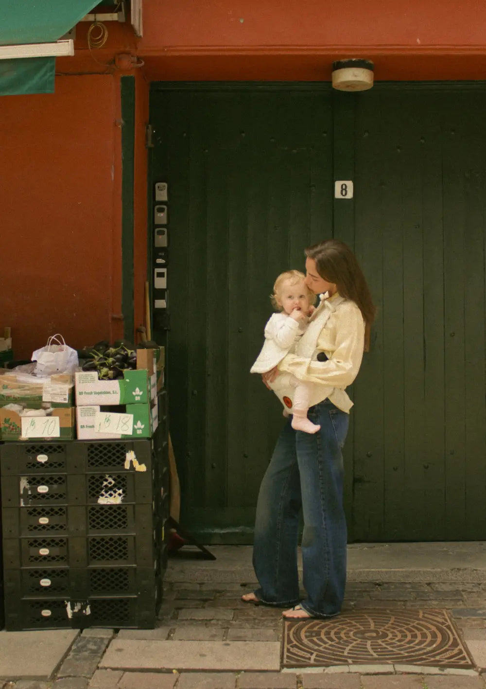 Eine Mutter steht vor einer grünen Einfahrt und trägt ihrer Kind in der Moova Babytrage 'Balance' Soft Sand
