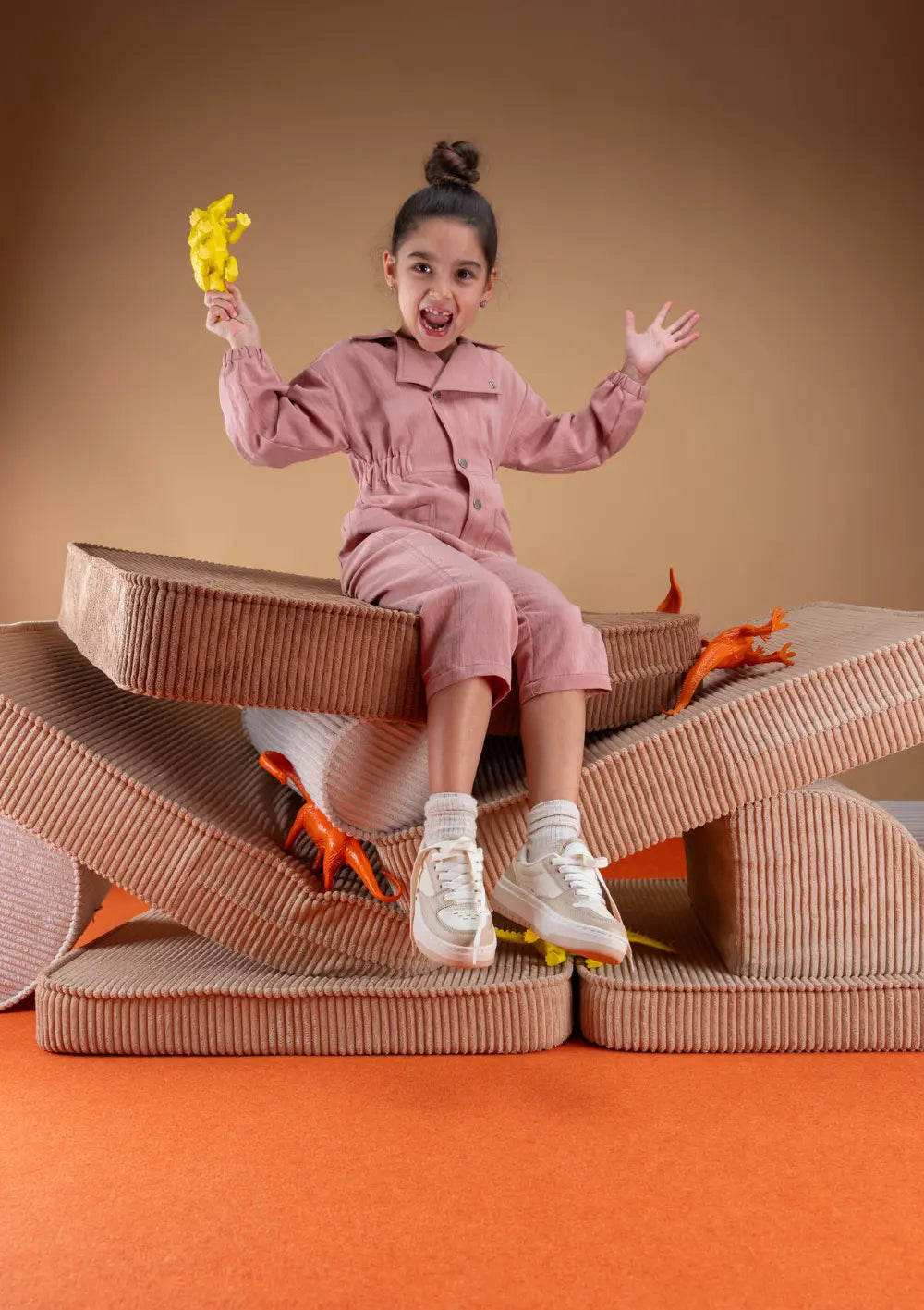 Ein Mädchen im rosa Overalls sitzt jubelnd auf einer aufeinandergestapelten Spiellandschaft aus den Bausteinen des modularen Wigiwama Kindersofas Cord Toffee und hält eine Spielzeugfigur in der Hand.