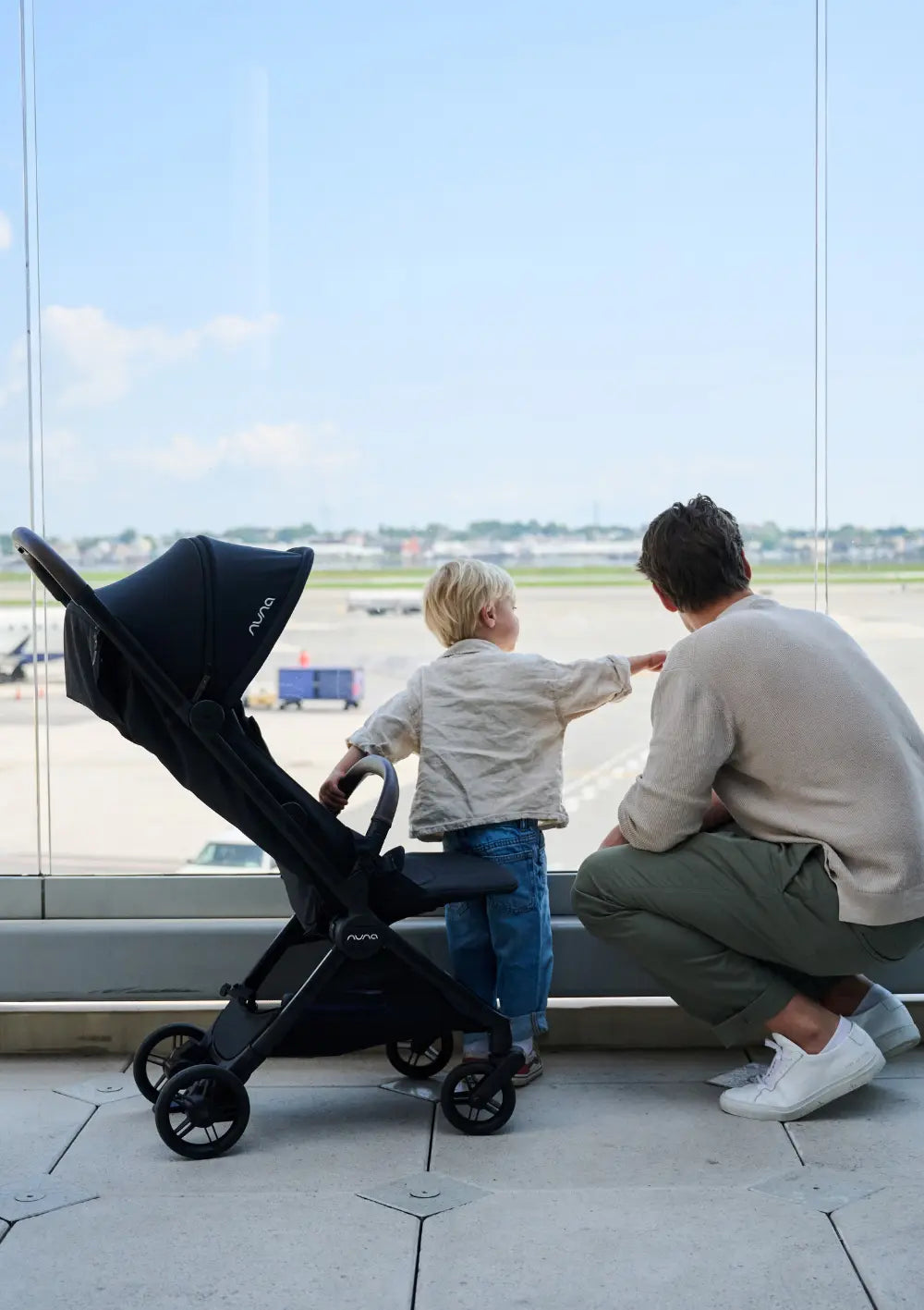 Ein Vater und sein Sohn stehen an einem großen Flughafenfenster und beobachten Flugzeuge auf dem Rollfeld. Links neben ihnen steht ein schwarzer Nuna VIAA cabn Buggy. Das Bild vermittelt Reiselust und die Rolle des Buggys als praktischer Reisebegleiter im Terminal.