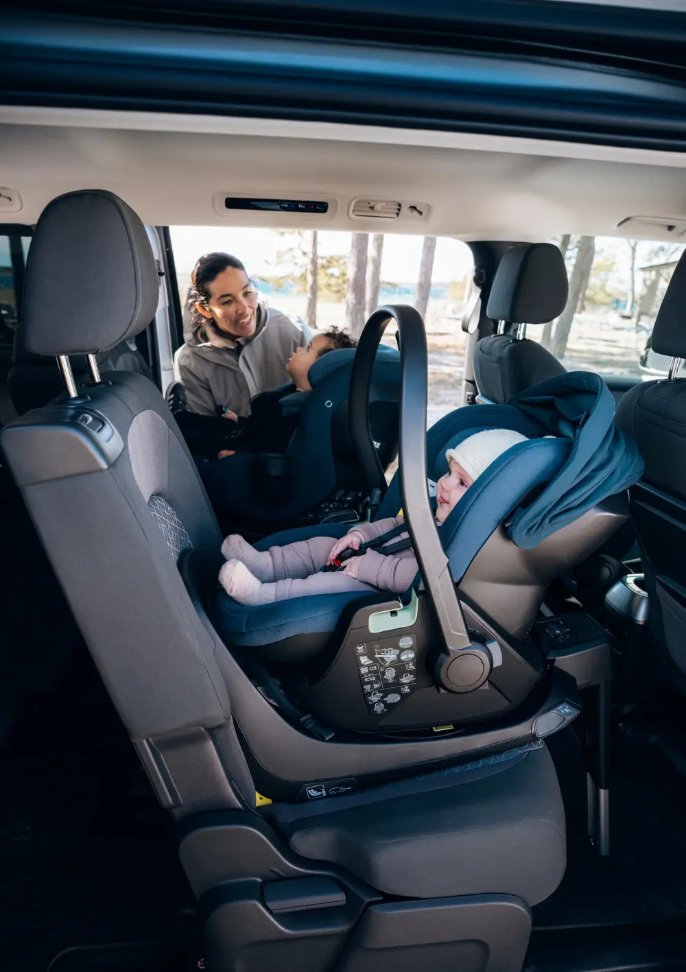 Eine Mutter schnallt ihre Kinder im Auto an. Das Baby sitzt in der Thule Babyschale 'Maple Connect' Darkest Blue, das Kleinkind im Thule Kindersitz 'Elm Connect 360' Darkest Blue.