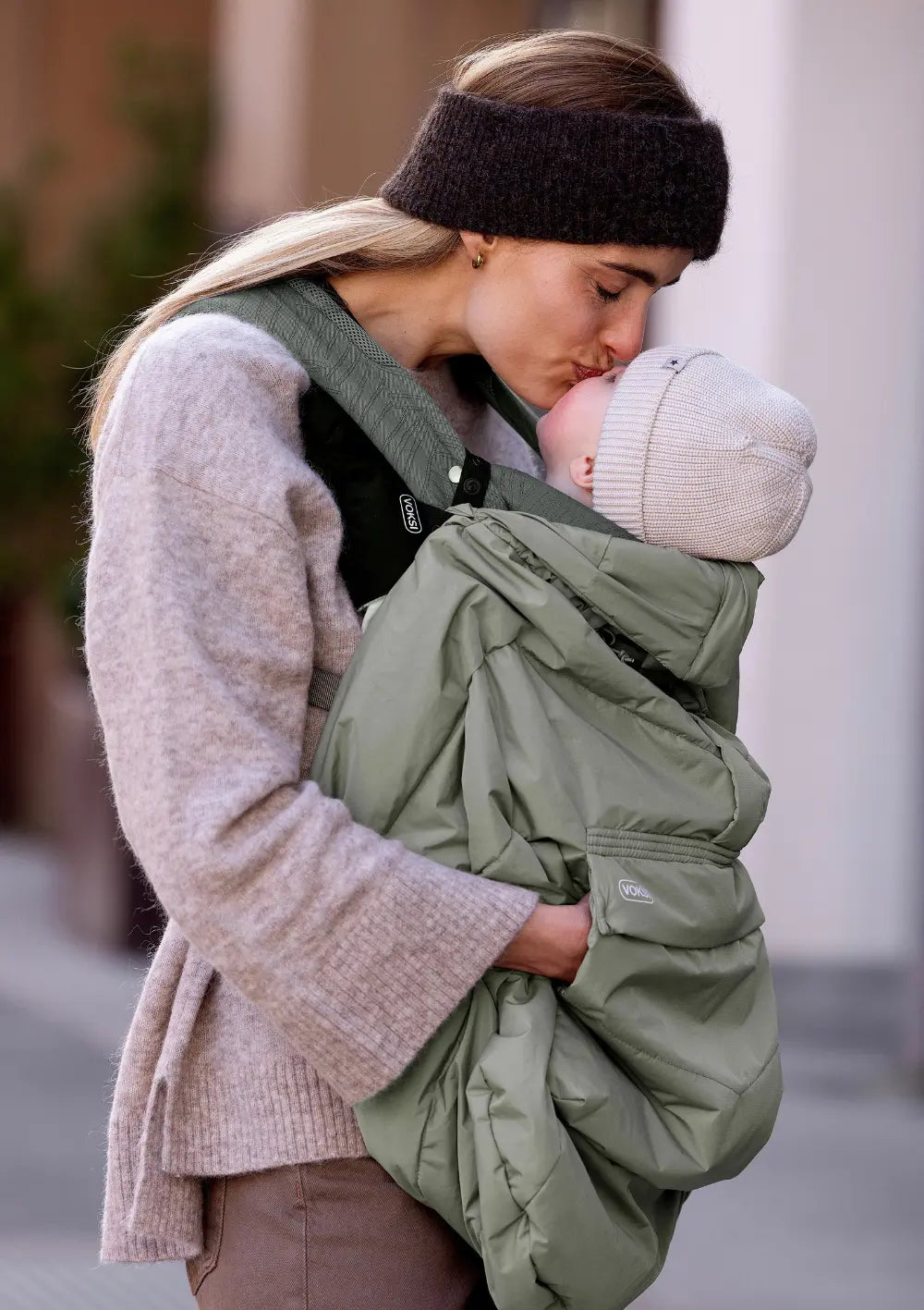 Mutter küsst ihr Baby, das in der Babytrage Huddle von Voksi sitzt, geschützt durch das Cover für Babytrage Meadow Green