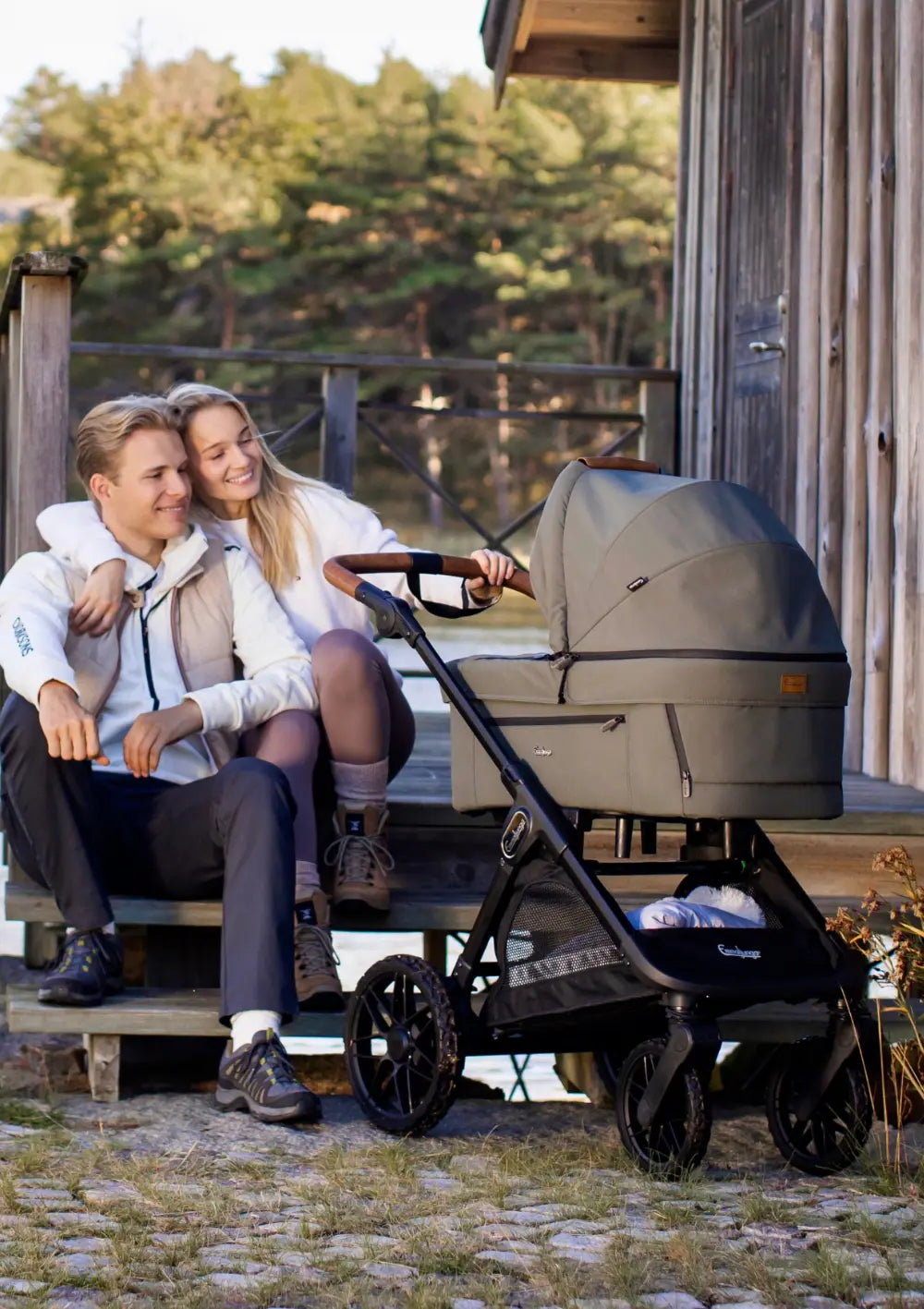 Ein Paar sitzt entspannt auf den Holzstufen einer Hütte und schaut lächelnd auf den olivenfarbenen Kombikinderwagen Emmaljunga SENTO PRO Outdoor Green.