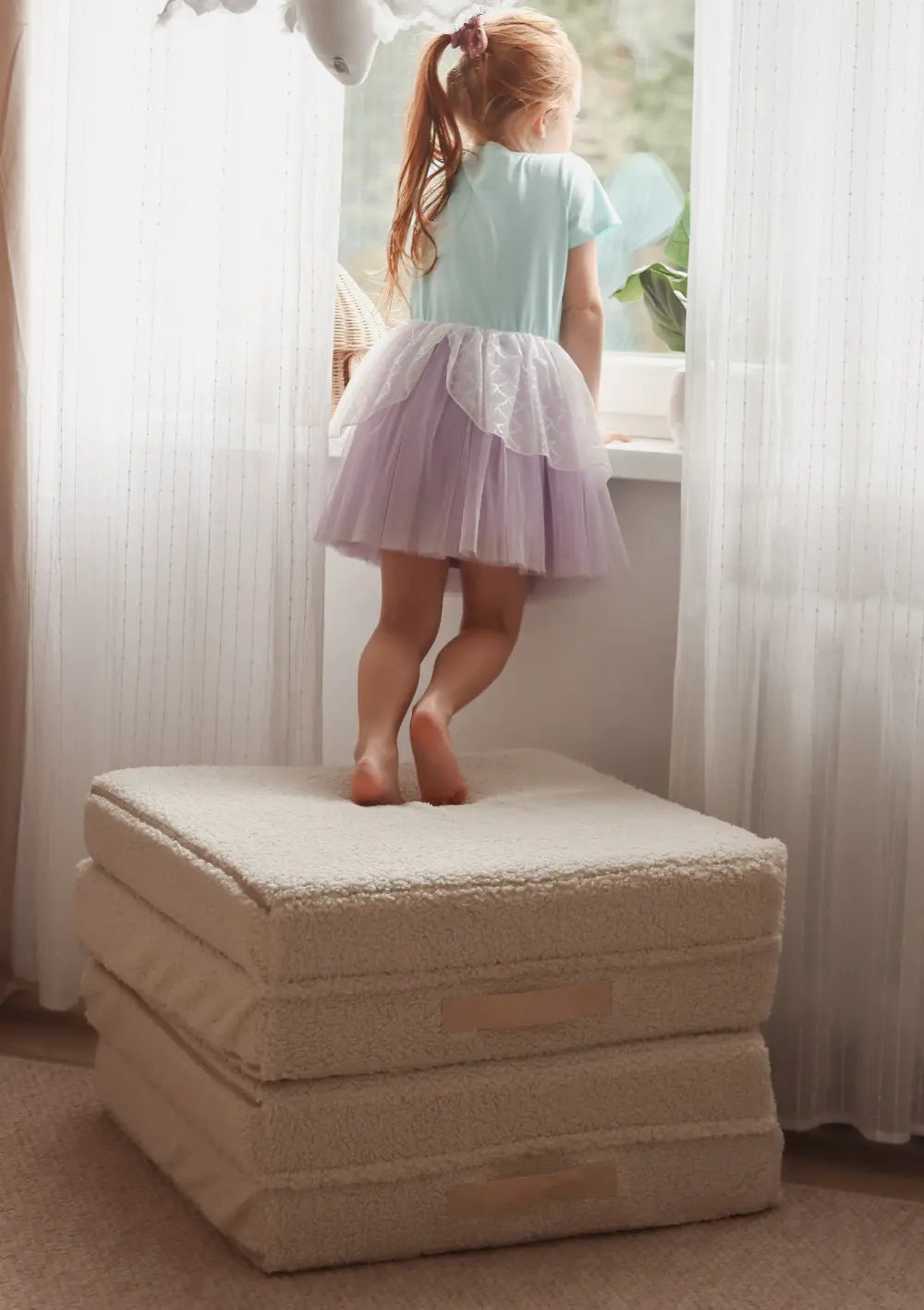 Mädchen steht auf den Polsterelementen des Funzy Mini Spielsofas Teddy Beige und schaut aus dem Fenster