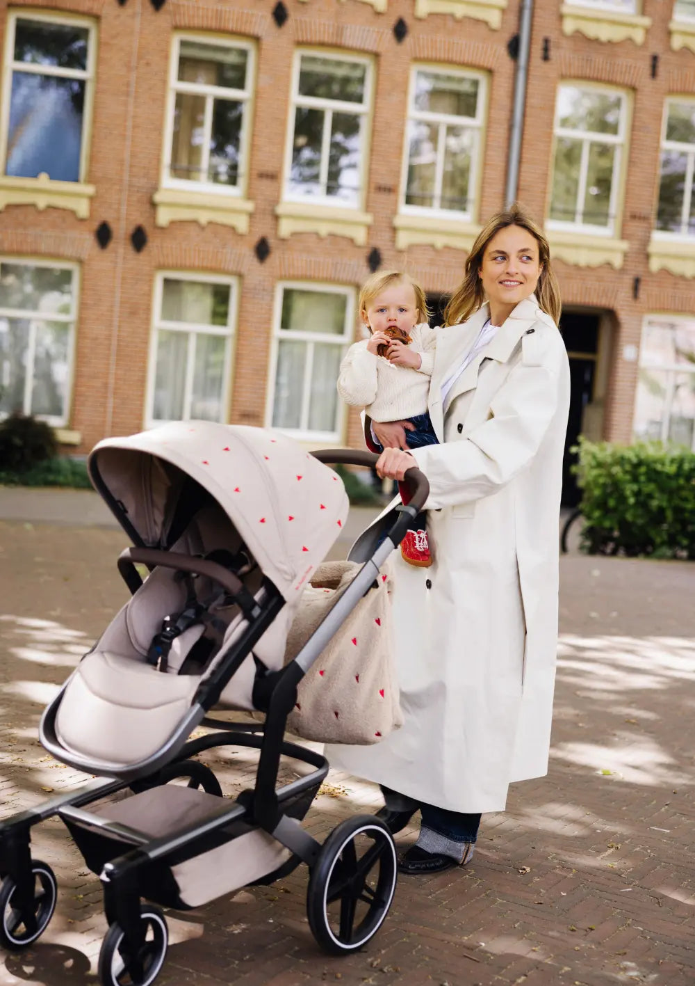 Eine Frau im hellen Mantelschiebt den Buggy des Joolz Kinderwagen-Set Day5 x Studio Noos Sandy Taupe mit Herzen-Muster auf dem Sonnendach durch die Stadt, während sie ein Kleinkind auf dem Arm hält.