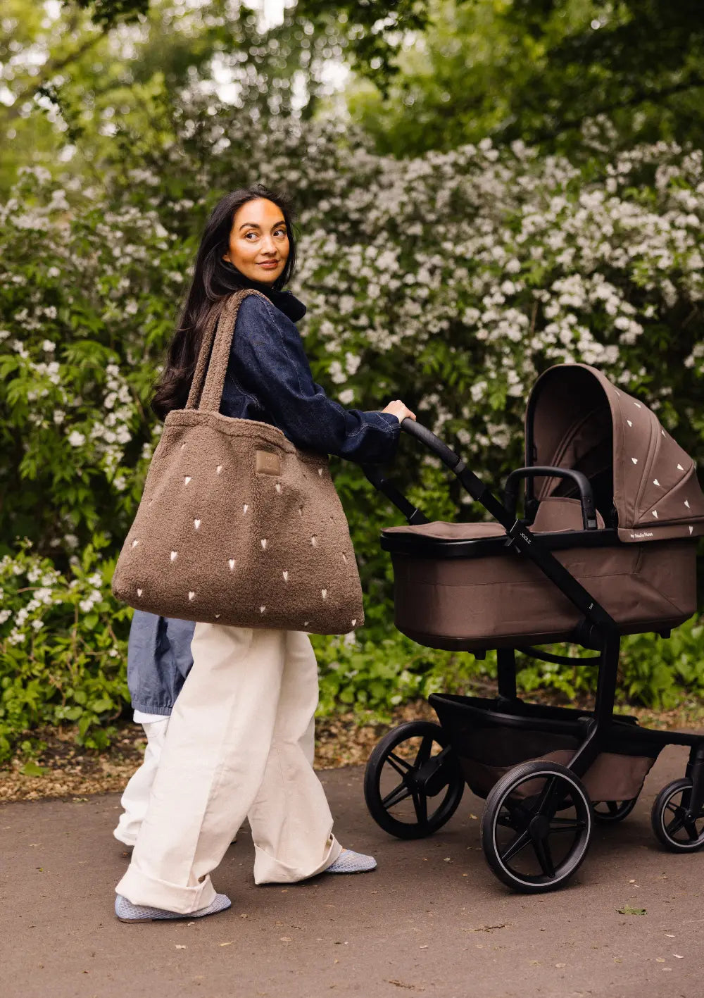 Eine Frau im Park schiebt den braunen Kombikinderwagen aus dem Joolz Kinderwagen-Set Day5 x Studio Noos mit Herzen und trägt die passende flauschige Studio Noos Tasche über der Schulter.