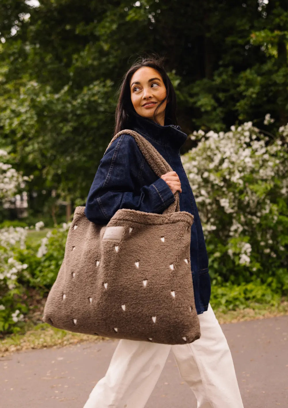 Eine Frau im Jeanshemd spaziert im Freien und trägt die braune Studio Noos Teddy-Shopper-Tasche mit kleinen weißen Herzen über der Schulter.