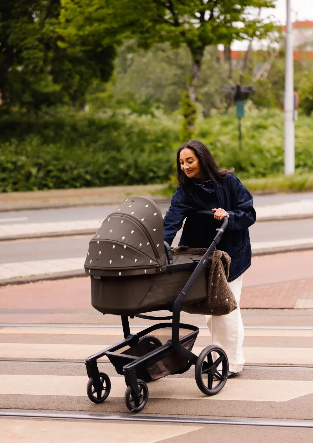Eine Mutter überquert eine Straße und schiebt dabei den braunen Kombikinderwagen des Joolz Kinderwagen-Set Day5 x Studio Noos mit Herzen auf dem Sonnendach