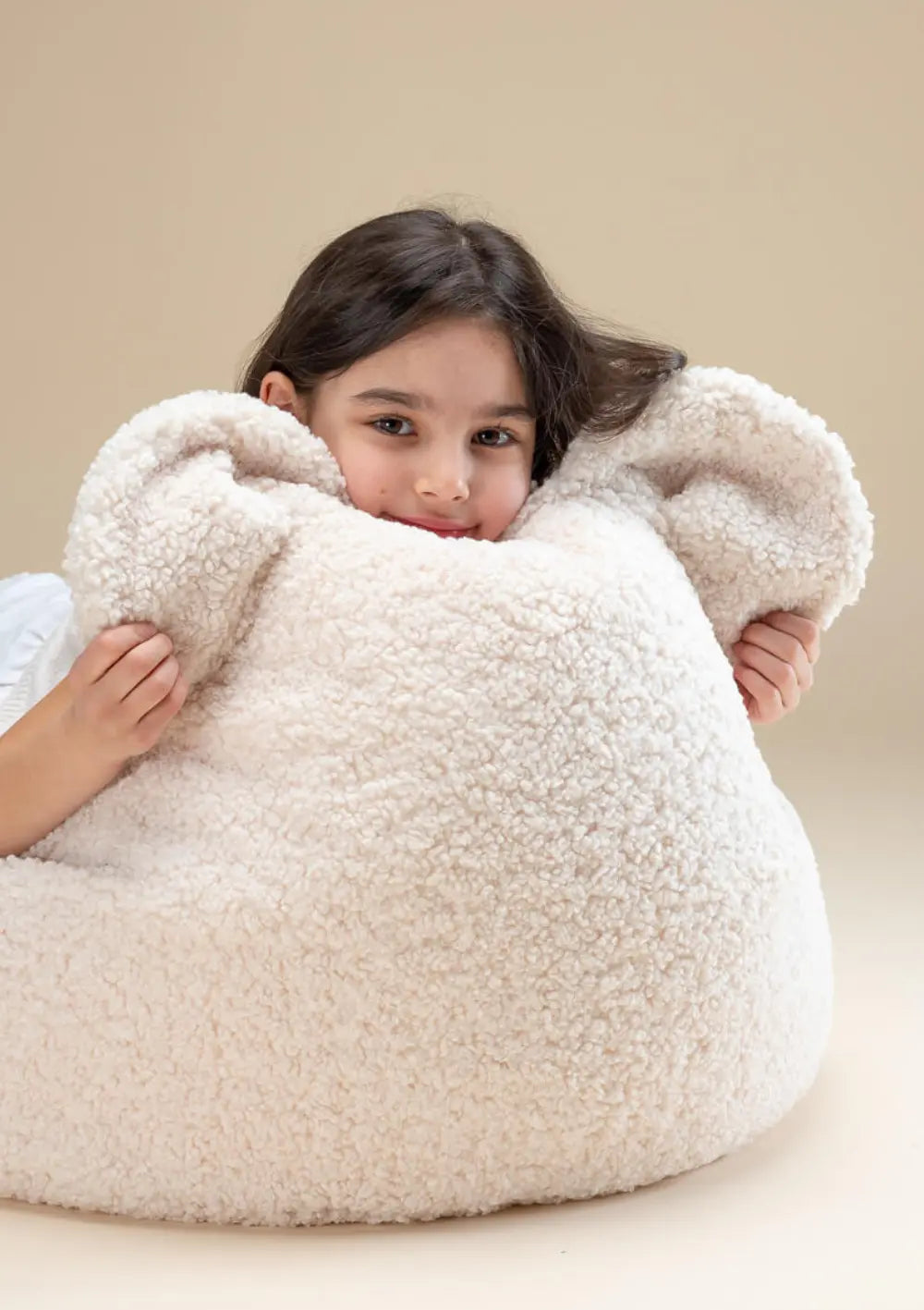 Ein Mädchen schaut lächelnd hinter der Rückenlehne mit Bärenohren des cremeweißen Wigiwama Bären Sitzsack Cream Teddy White hervor.