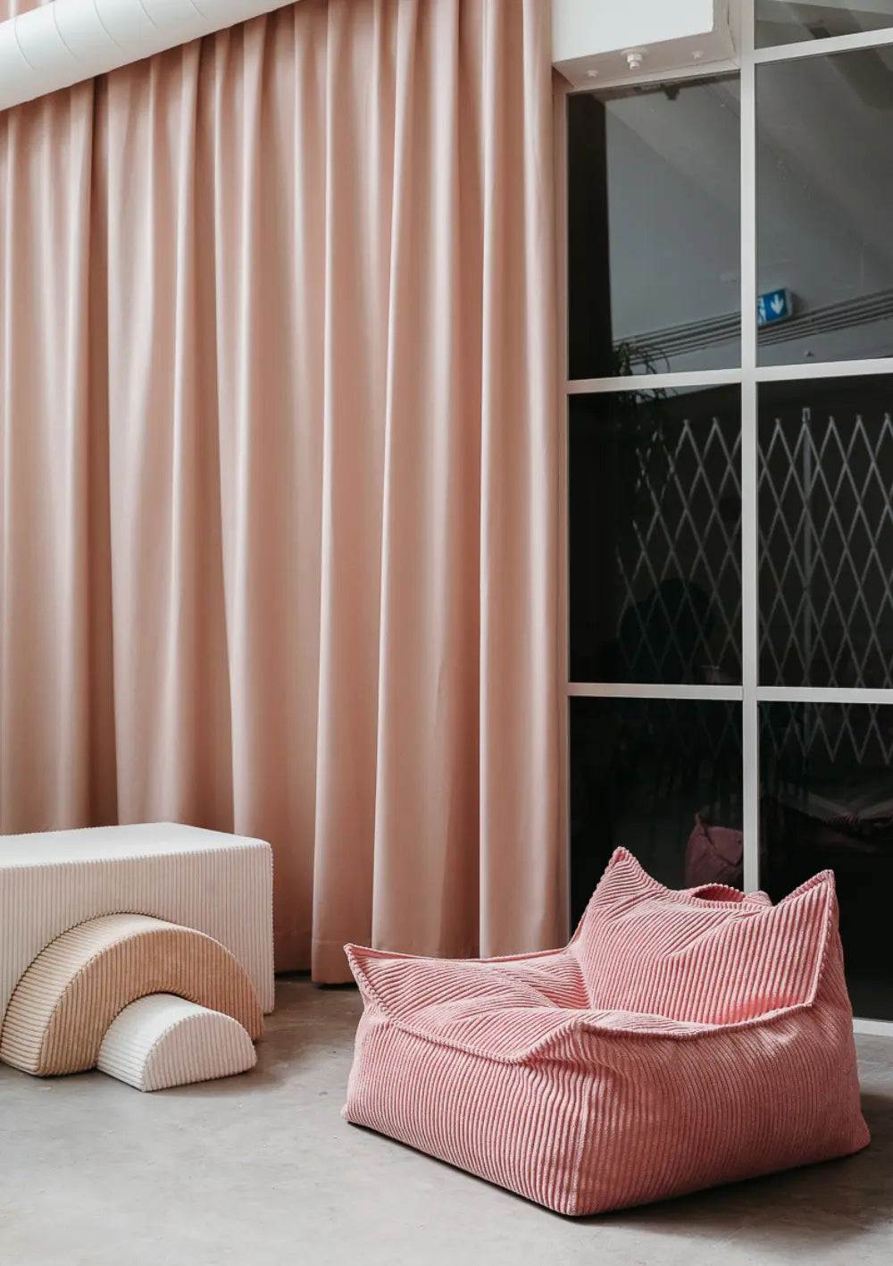 Der rosafarbene Wigiwama Sitzsack Cord Pink Mousse aus weichem Breitcord steht freistehend vor einem Vorhang und großen Fensterelementen in einem stilvoll eingerichteten Raum.