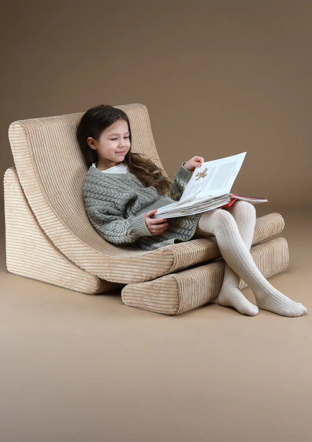 Ein Mädchen im grauen Strickkleid sitzt entspannt im hellbraunen Wigiwama Kindersessel Moon Chair Cord Brown Sugar und blättert in einem Buch.