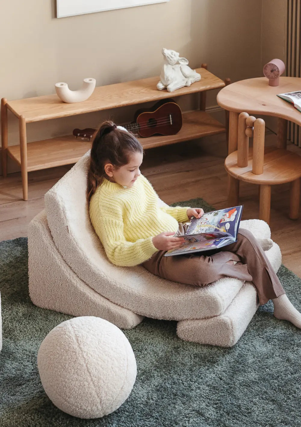 Ein Mädchen im gelben Pullover sitzt im cremeweißen Wigiwama Kindersessel Moon Chair Teddy Cream White aus kuschligem Plüsch und liest ein Bilderbuch.