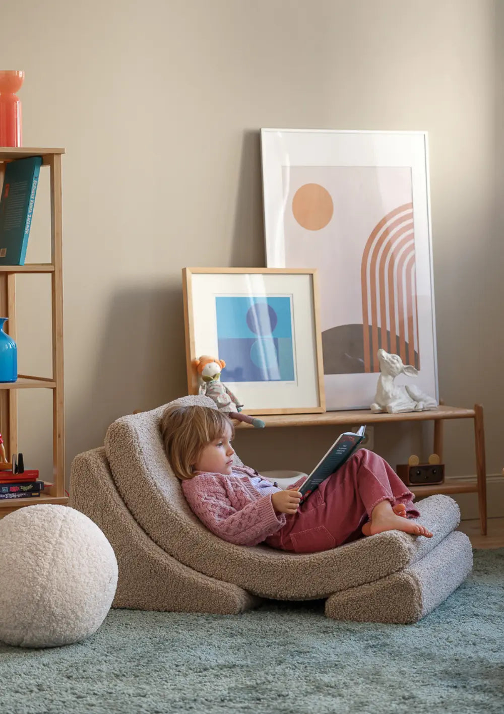 Ein Kind liegt entspannt im beigefarbenen Wigiwama Kindersessel Moon Chair Teddy Biscuit aus kuschligem Teddy-Stoff und liest aufmerksam ein Buch.