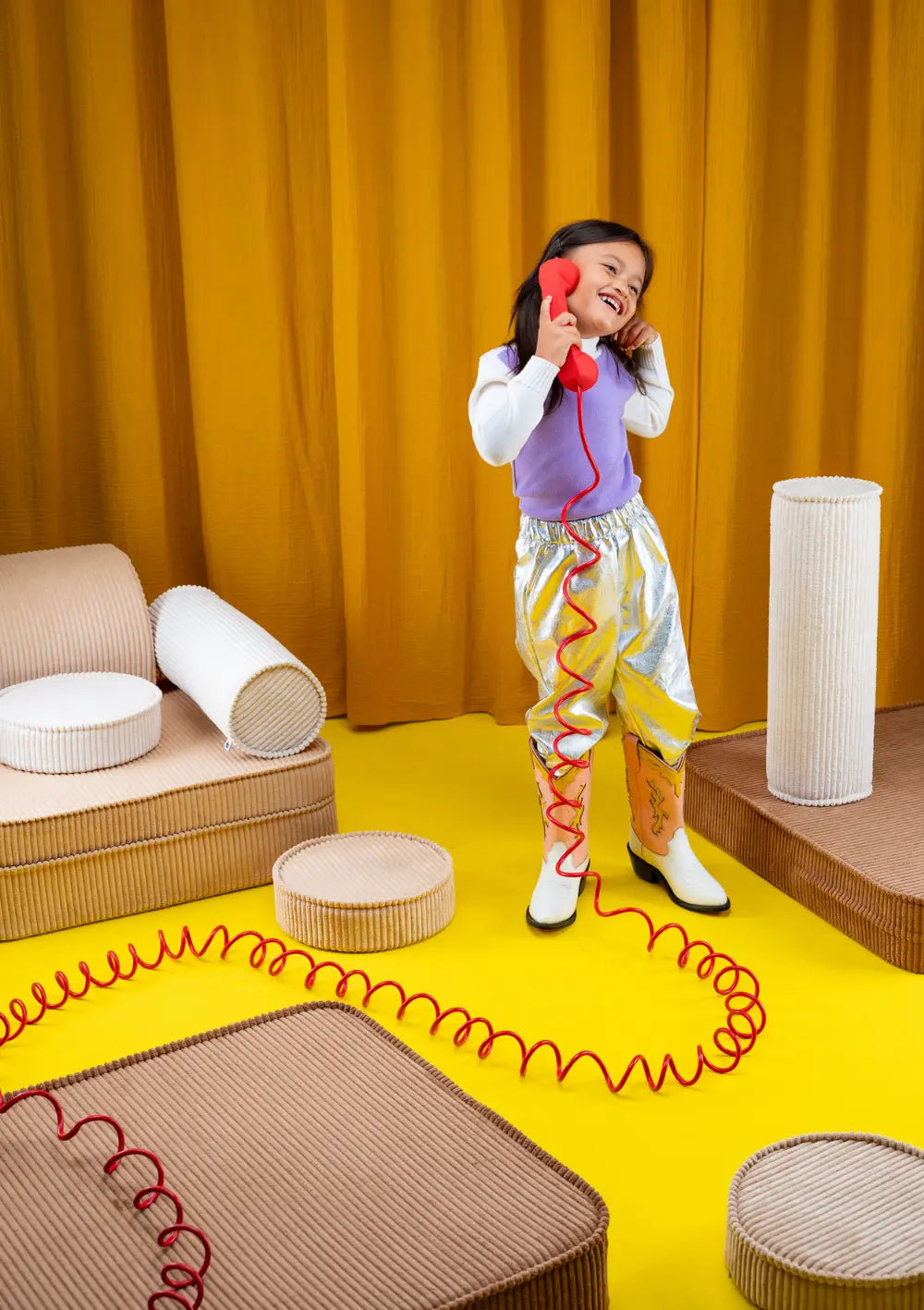 Ein Mädchen im lila Oberteil und Glanzhose telefoniert fröhlich mit einem roten Spieltelefon, umgeben von Bausteinen des modularen Wigiwama Kindersofas Cord Toffee.