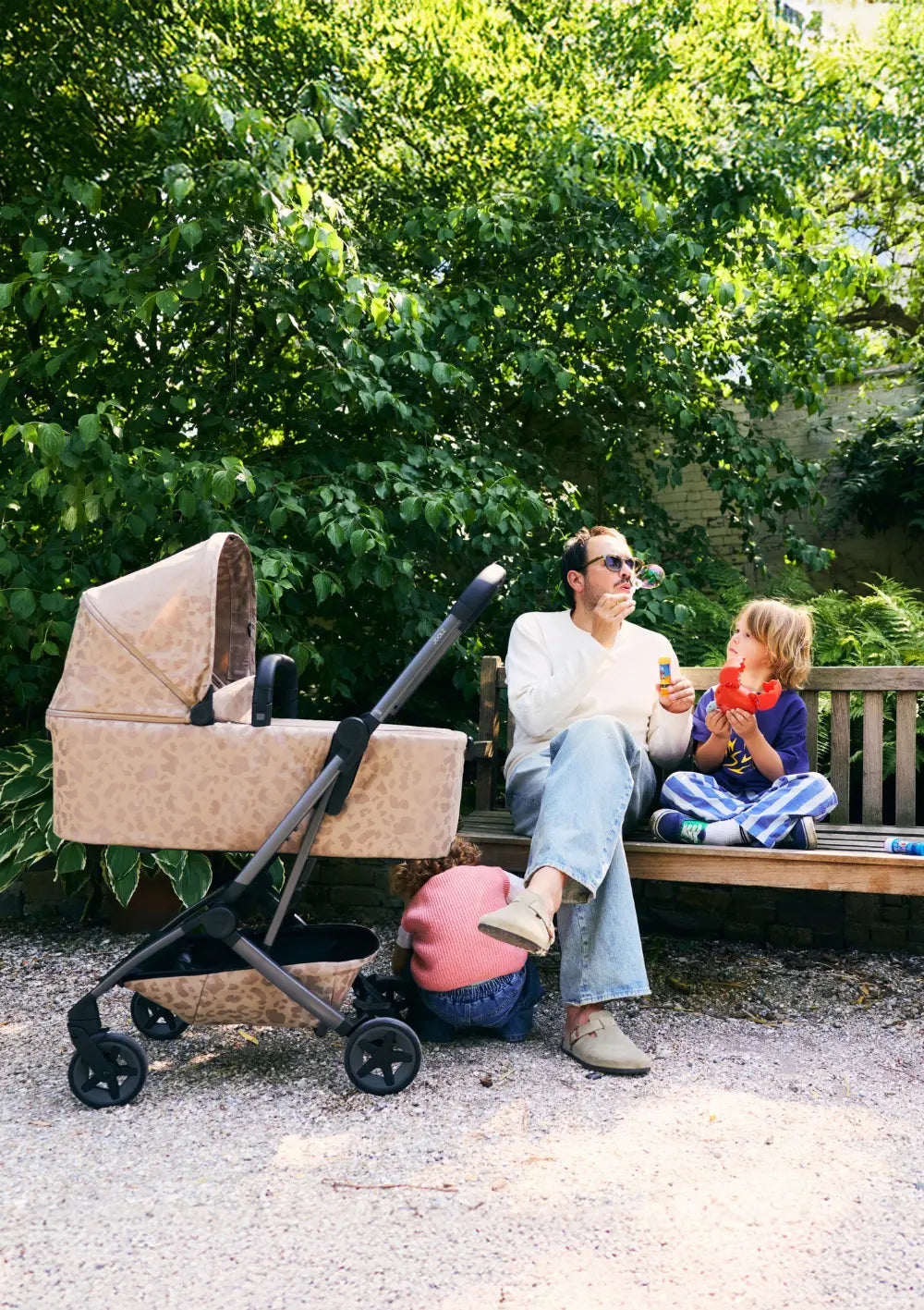 Ein Vater bläst auf einer Parkbank Seifenblasen für seine Kinder, während der  beigefarbene Joolz Kinderwagen Aer2 Chic Renaissance Sandy Taupe mit passender Babywanne  daneben steht.